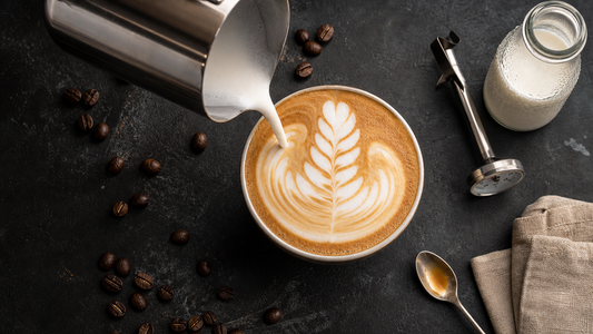 Steam pitcher pouring glossy microfoam into a flat white with a rosetta, milk bottle and thermometer on a dark surface
